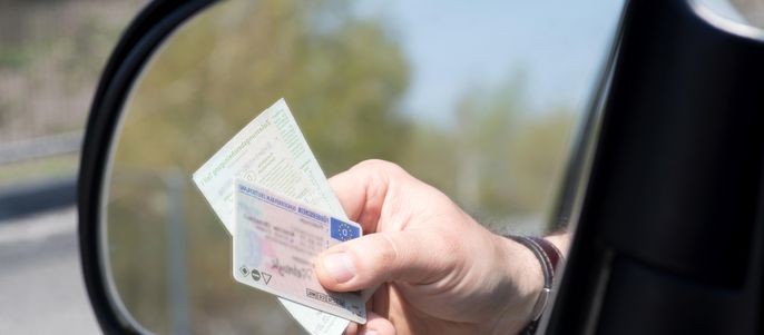Eine Hand, die einen Führerschein hält, wobei sich der Führerschein im Seitenspiegel des Autos spiegelt.