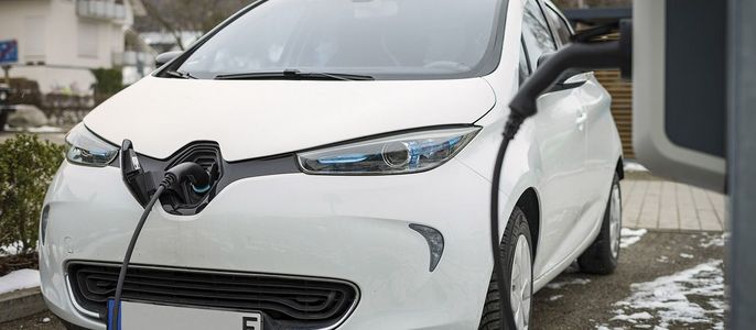 Ein weißes Elektroauto wird an einer Ladestation aufgeladen, mit einem Haus und einer verschneiten Umgebung im Hintergrund.