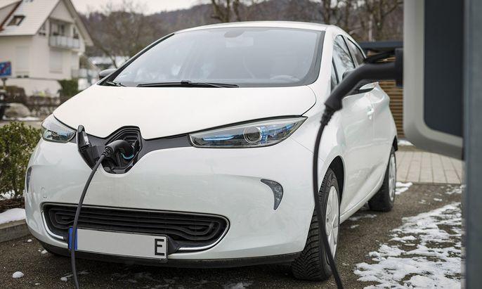 Ein weißes Elektroauto wird an einer Ladestation aufgeladen, mit einem Haus und einer verschneiten Umgebung im Hintergrund.