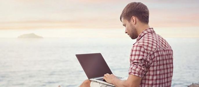 Ein Mann sitzt am Meer und benutzt einen Laptop.