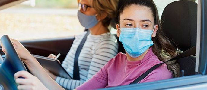 Zwei Frauen in einem Auto, beide mit Gesichtsmasken, wobei die Frau vorne das Lenkrad hält.