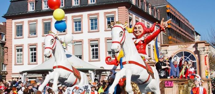 Der Mainzer Rosenmontagsumzug mit einem großen Wagen mit zwei weißen Pferden, einem Clown und verschiedenen bunten Elementen vor der Kulisse historischer Gebäude.