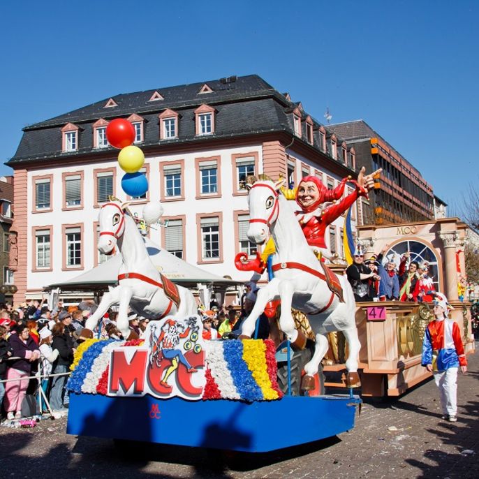 Der Mainzer Rosenmontagsumzug mit einem großen Wagen mit zwei weißen Pferden, einem Clown und verschiedenen bunten Elementen vor der Kulisse historischer Gebäude.